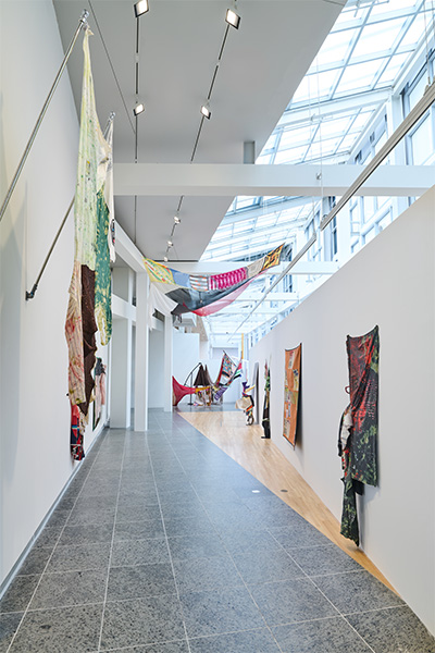 A gallery space featuring an installation of colorful fabric sculptures and paintings hanging from the walls and ceiling.