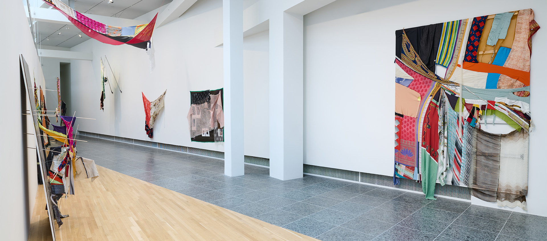 A gallery space featuring an installation of colorful fabric sculptures and paintings hanging from the walls and ceiling.