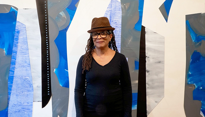 Nanette Carter with her artwork. She has light-brown skin, dark hair, and wears a fedora, glasses, a black outfit, a gold cuff, and rings.
