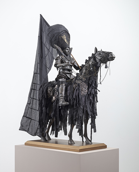 A grayish black statue of a man in armor and a large headpiece. He rides a horse. The man carries a large black flag.