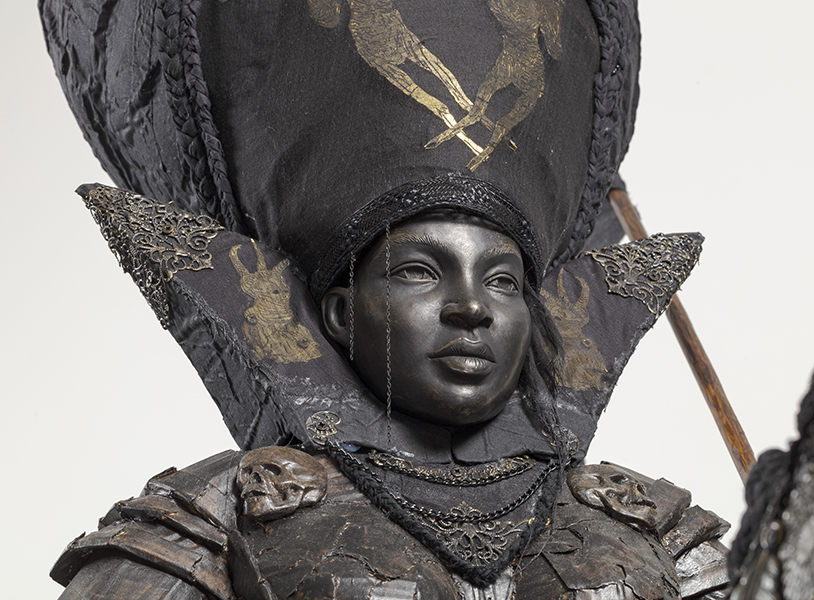 A grayish black bust of a regal-looking ambassador wearing a large headpiece embroidered in metals.
