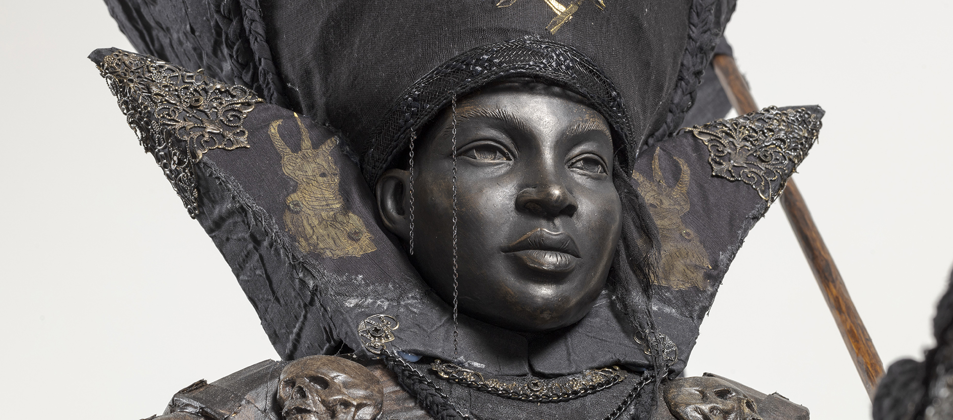 A grayish black bust of a regal-looking ambassador wearing a large headpiece embroidered in metals.