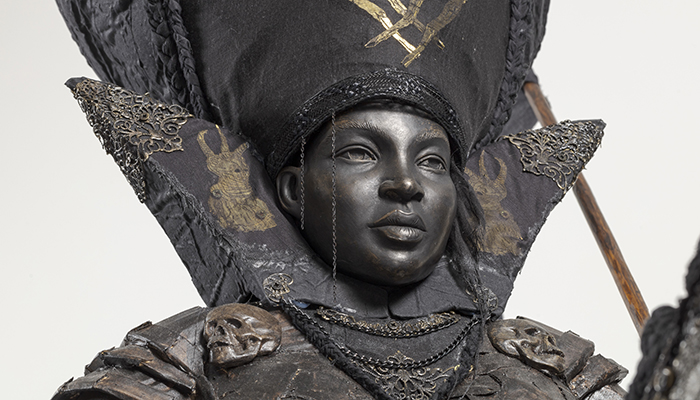 A grayish black bust of a regal-looking ambassador wearing a large headpiece embroidered in metals.