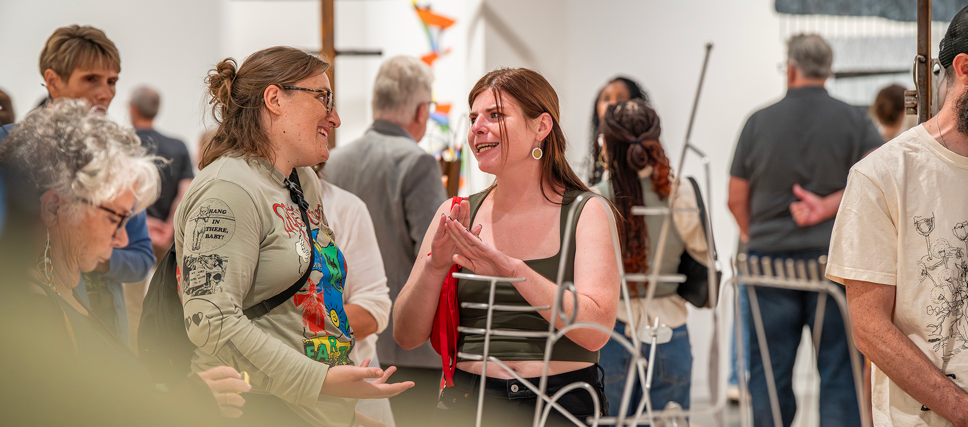 Two people stand behind a wire sculpture conversing. Additional people around them look at artwork in the gallery.  