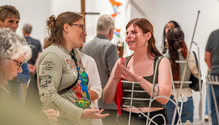 Two people stand behind a wire sculpture conversing. Additional people around them look at artwork in the gallery.  