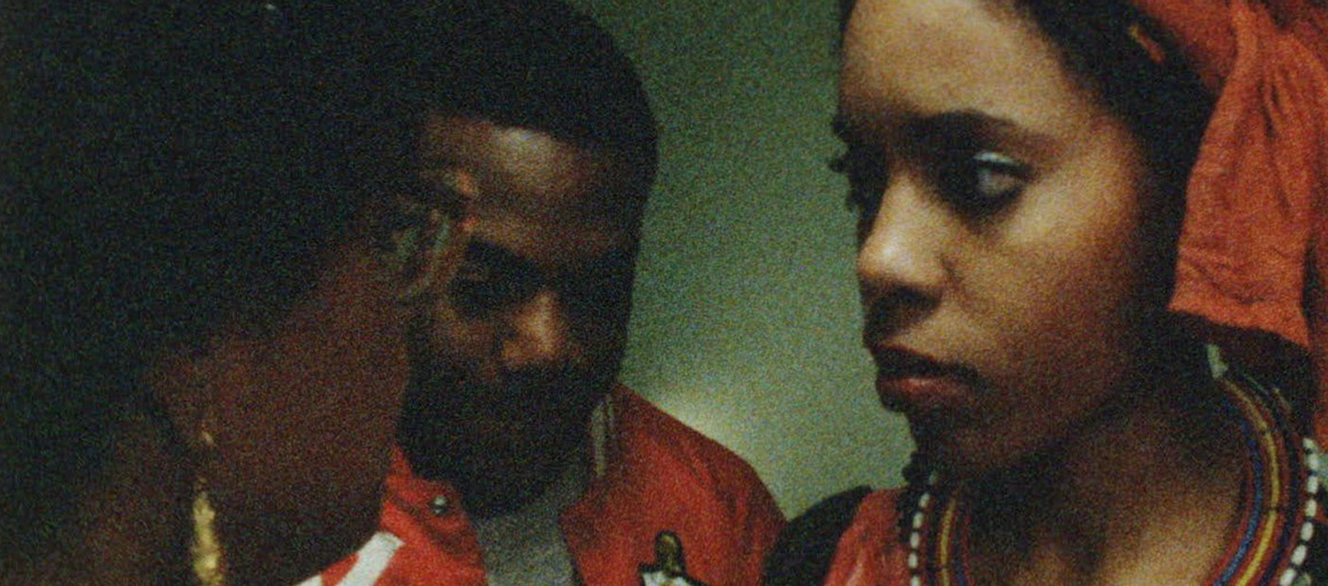 Three people are close together; a woman with a headwrap and beaded necklace looks at a person in glasses. A man stands in the background.