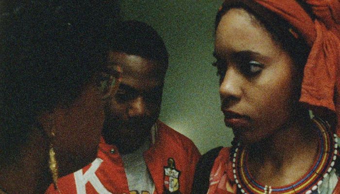 Three people are close together; a woman with a headwrap and beaded necklace looks at a person in glasses. A man stands in the background.