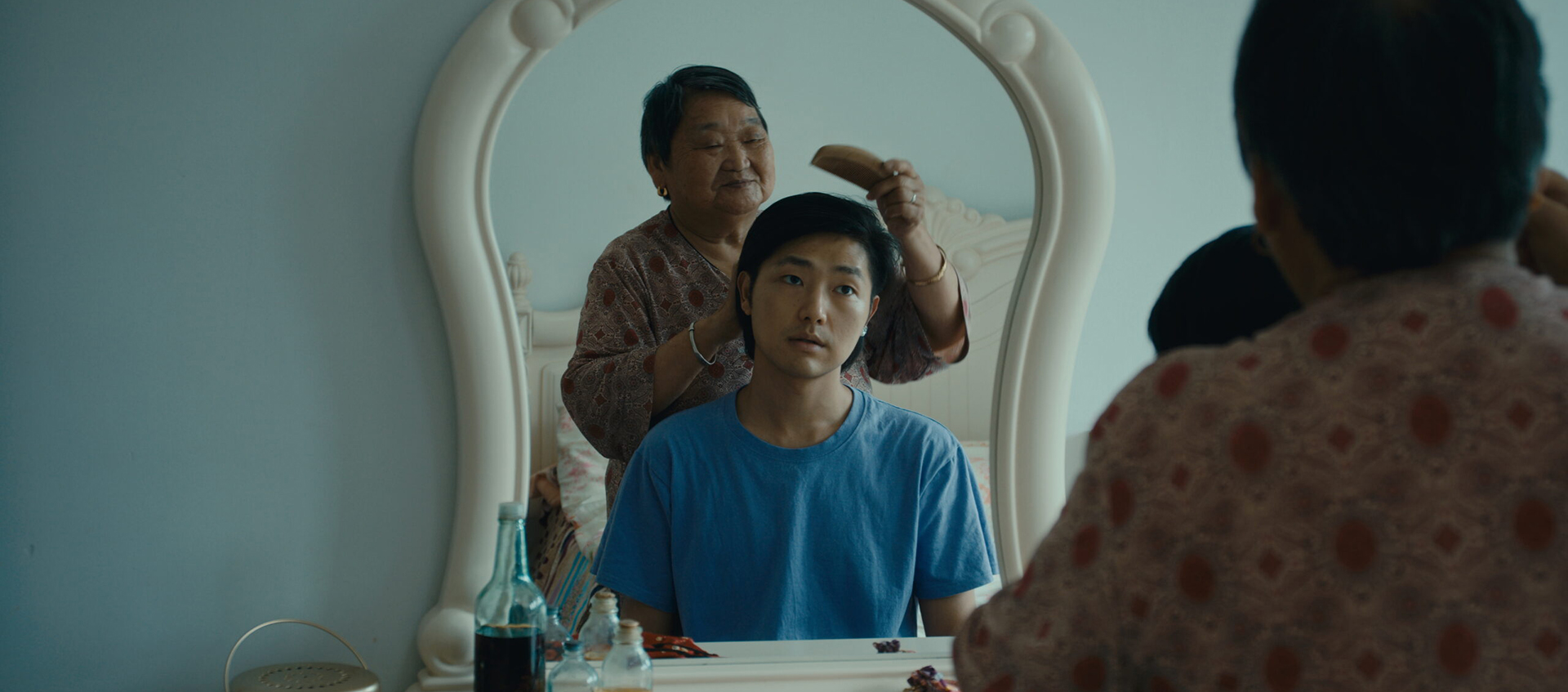 An older woman brushes a young person's hair in front of a mirror. The young person sits quietly in a blue shirt while the woman stands behind them. Various items are on the vanity.