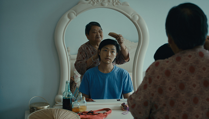 An older woman brushes a young person's hair in front of a mirror. The young person sits quietly in a blue shirt while the woman stands behind them. Various items are on the vanity.