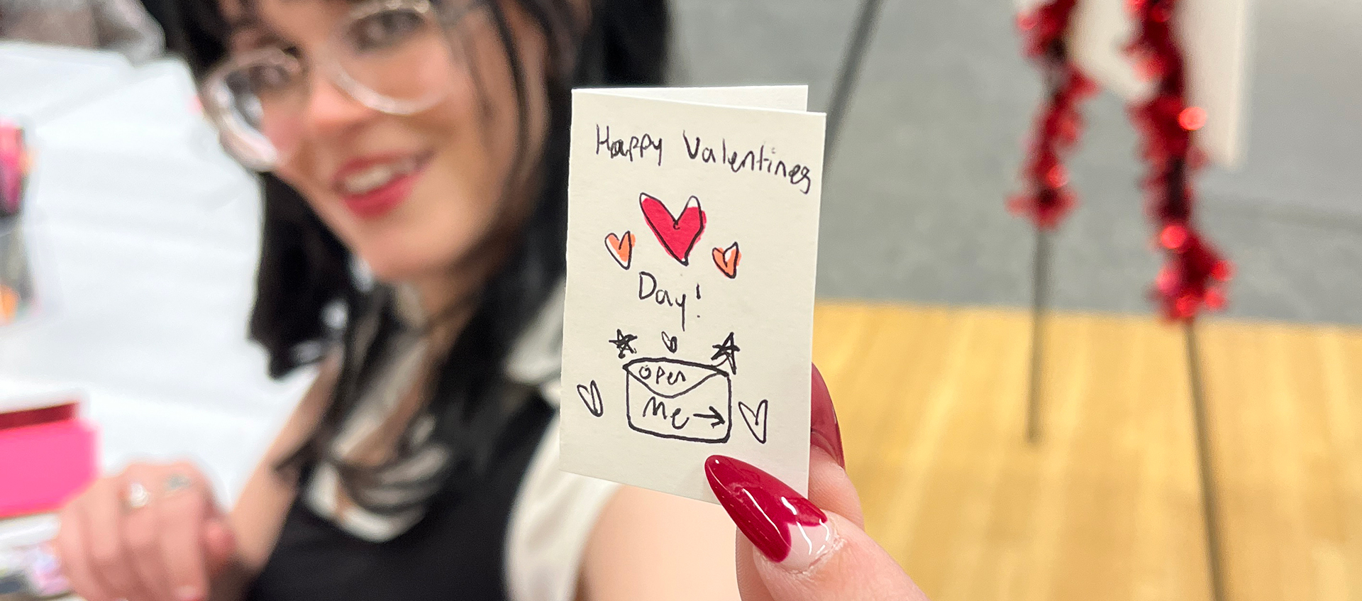 A young woman with fair skin and dark hair holds up a miniature card that reads Happy Valentine's Day! Open me.