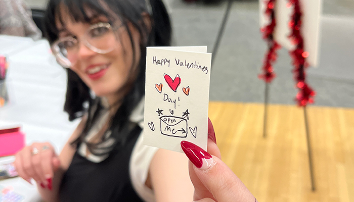 A young woman with fair skin and dark hair holds up a miniature card that reads Happy Valentine's Day! Open me.