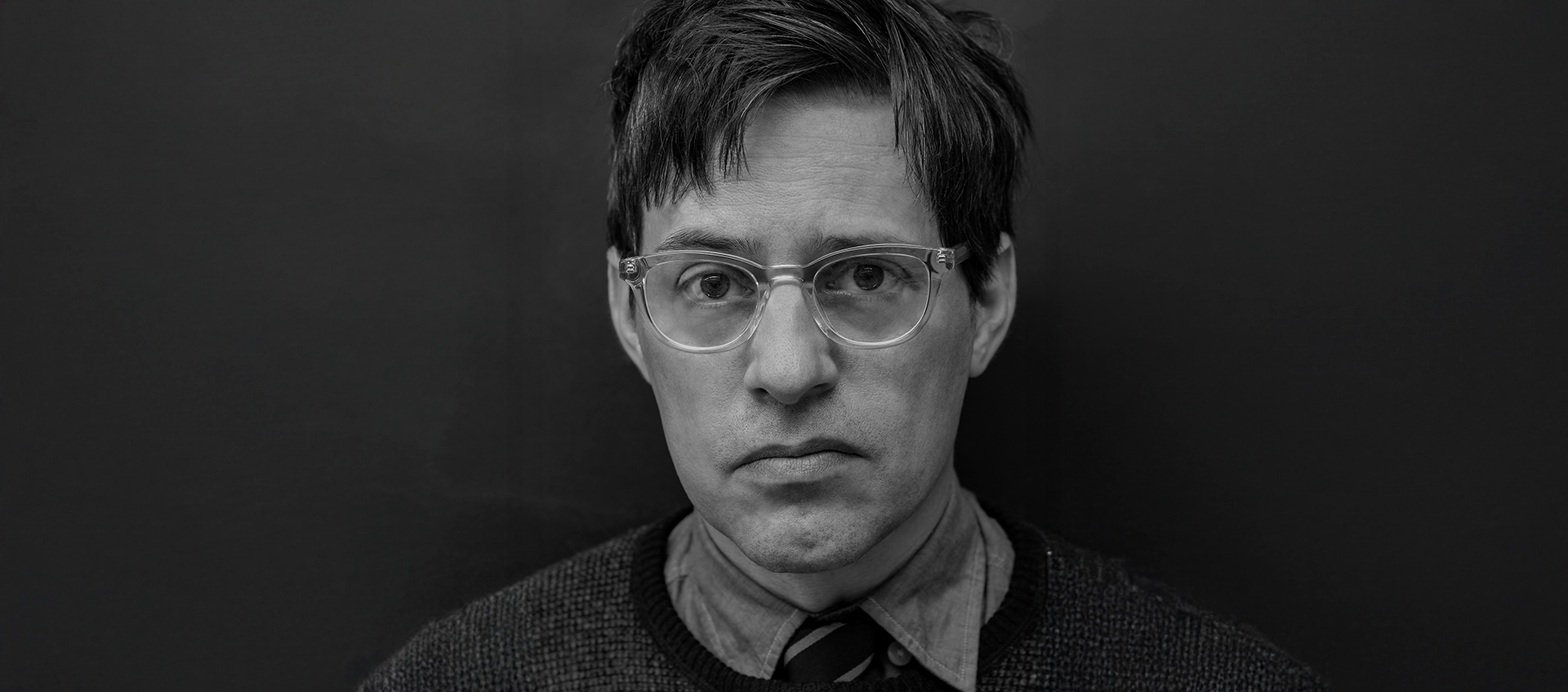 Portrait of Christian Patterson, who has light skin and short dark hair. He wears glasses, a collared shirt, and a sweater.