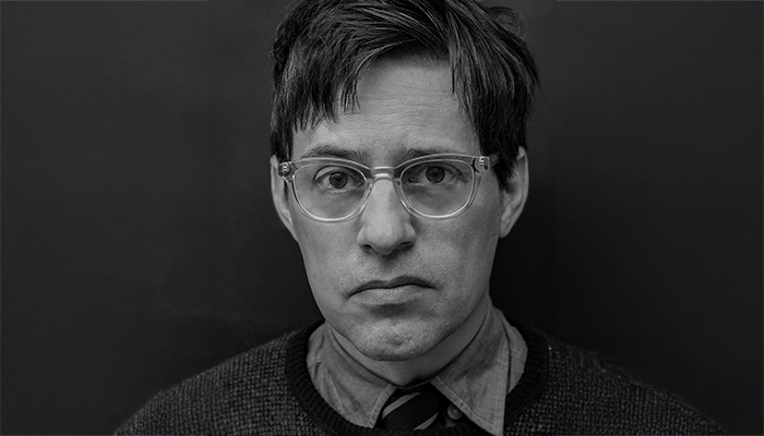 Portrait of Christian Patterson, who has light skin and short dark hair. He wears glasses, a collared shirt, and a sweater.