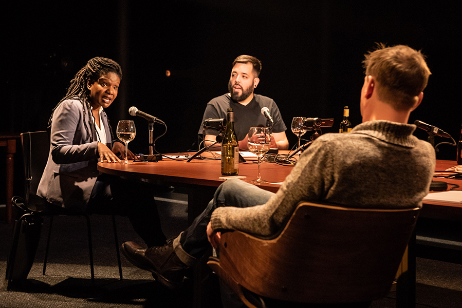 Two men and a woman sit at a long table. The woman is speaking with her hands pressed on the table.