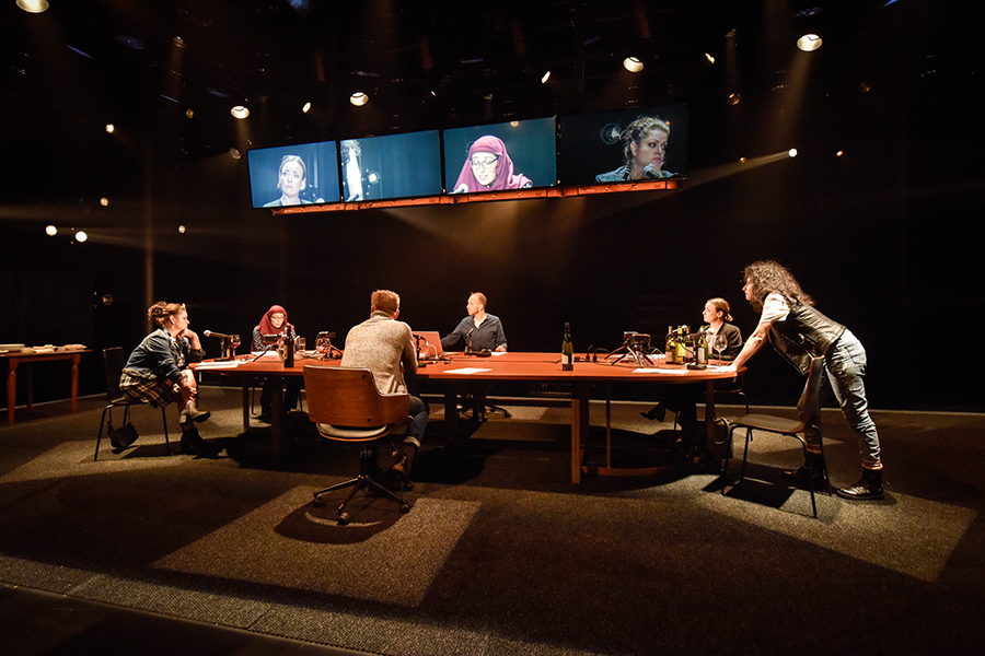 Five people sit and one leans on a table that has audio and video equipment. Monitors above show their faces.