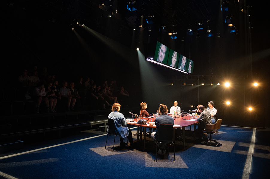 Six people sit at a long table with microphones and recording equipment on it. Monitors above them show their faces.