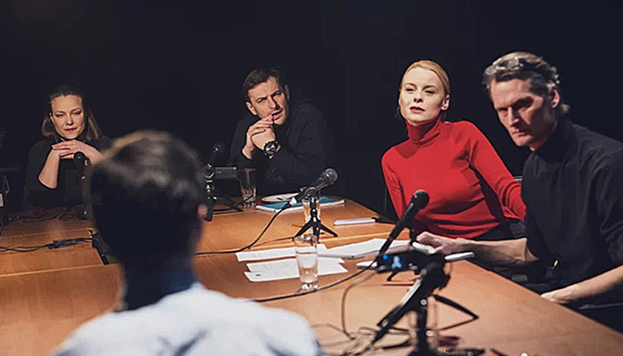 Five people sit around a table with video cameras and microphones on it. 
