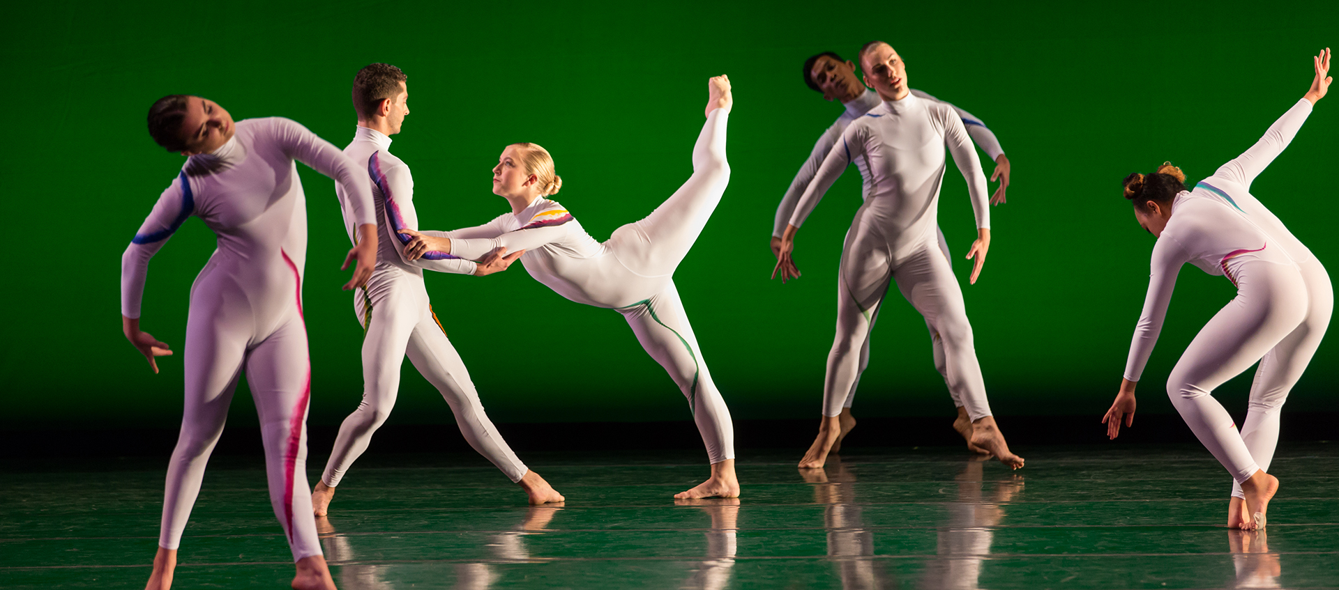 Six dancers in white bodysuits perform a contemporary dance routine on stage with a green background.