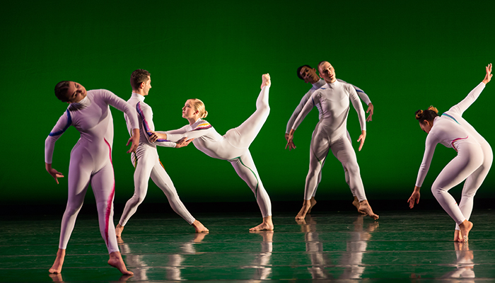 Six dancers in white bodysuits perform a contemporary dance routine on stage with a green background.