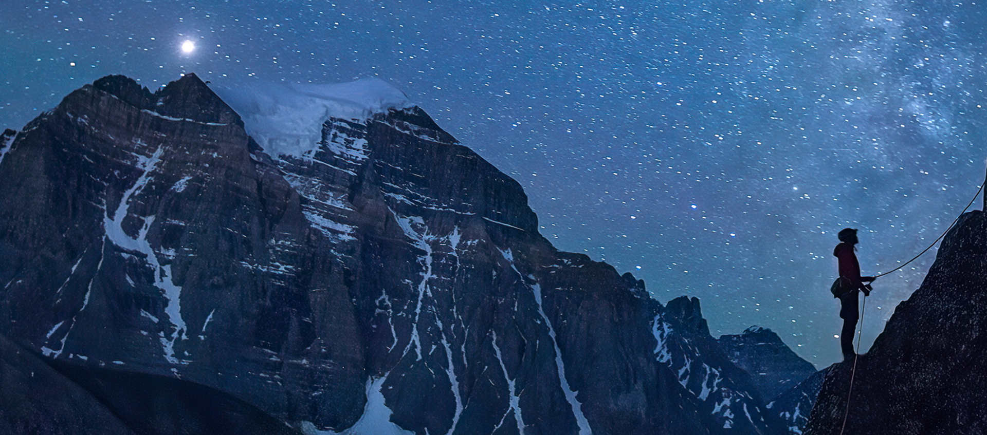 The silhouette of a rock climber holding a rope in front of a mountain and starry night sky.