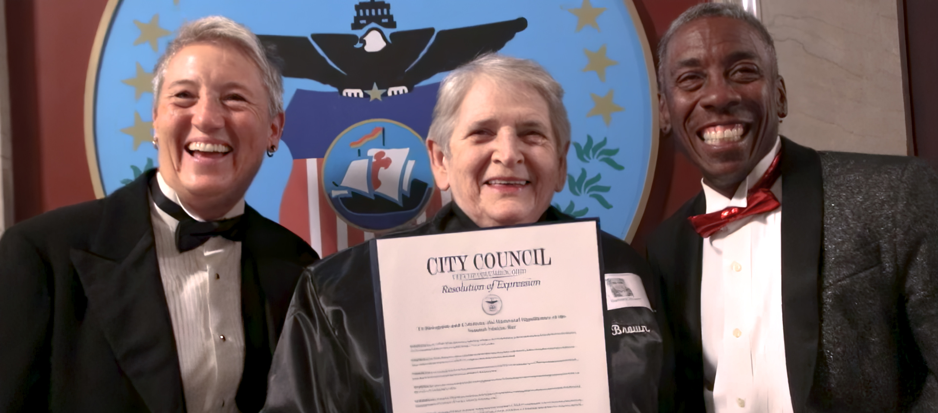 Three individuals stand in front of the US seal holding an official document.