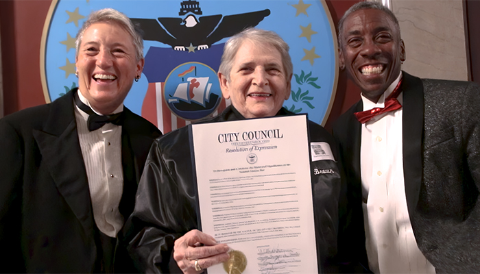 Three individuals stand in front of the US seal holding an official document.