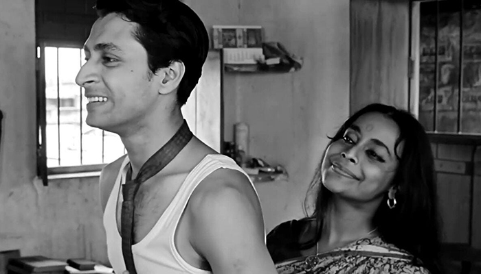 A man stands in an undershirt and tie while a woman in a sari stands behind him.