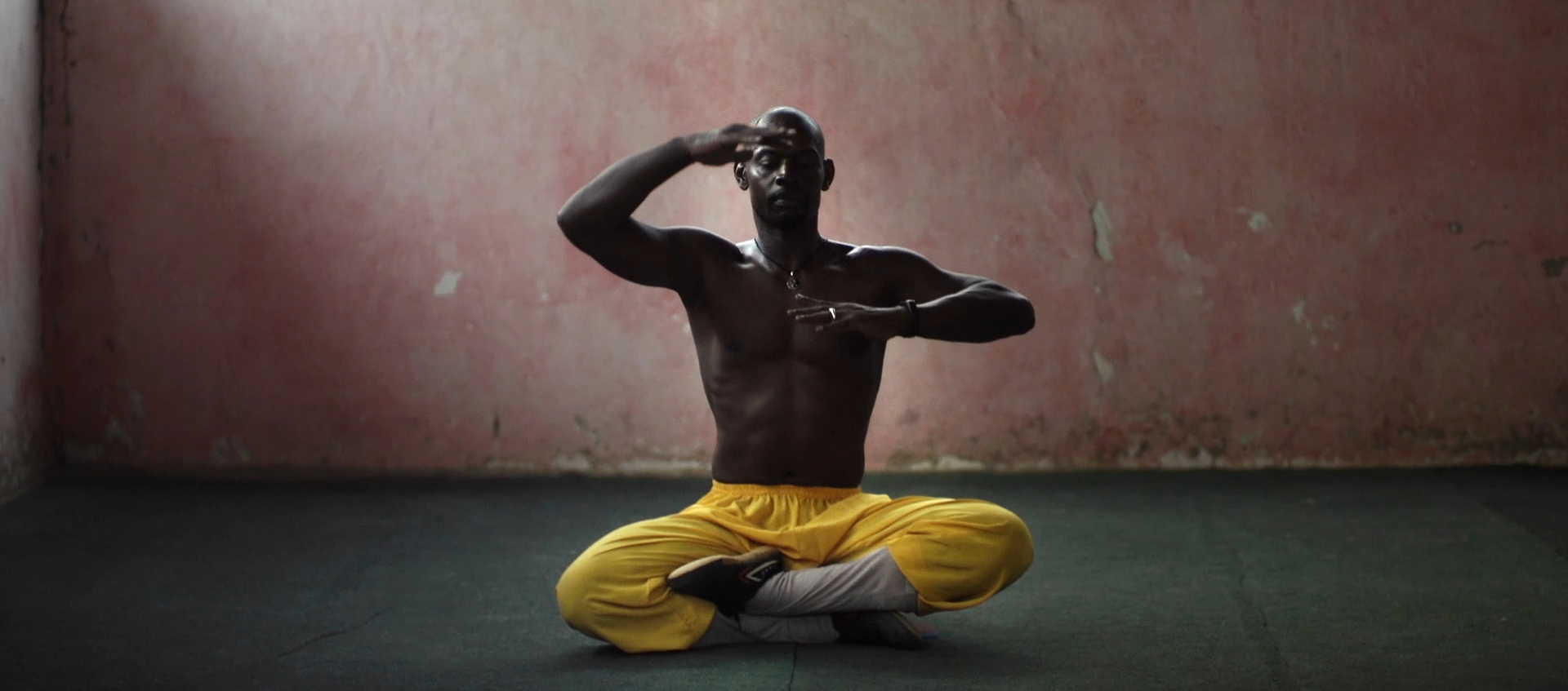 A man sits cross-legged on the floor with hands held in front of his face and chest.