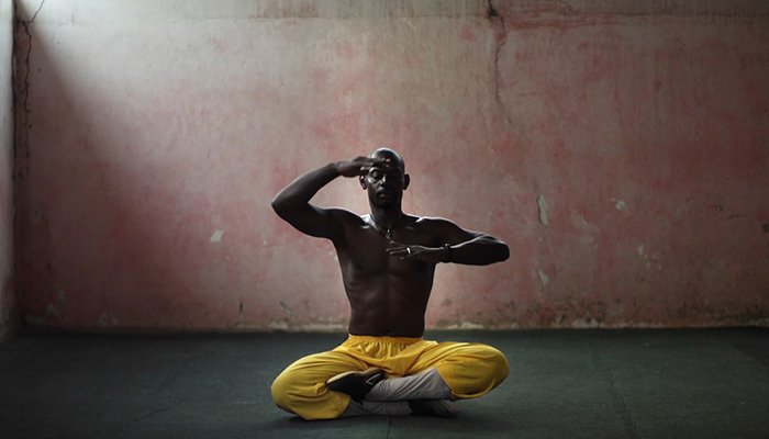 A man sits cross-legged on the floor with hands held in front of his face and chest.