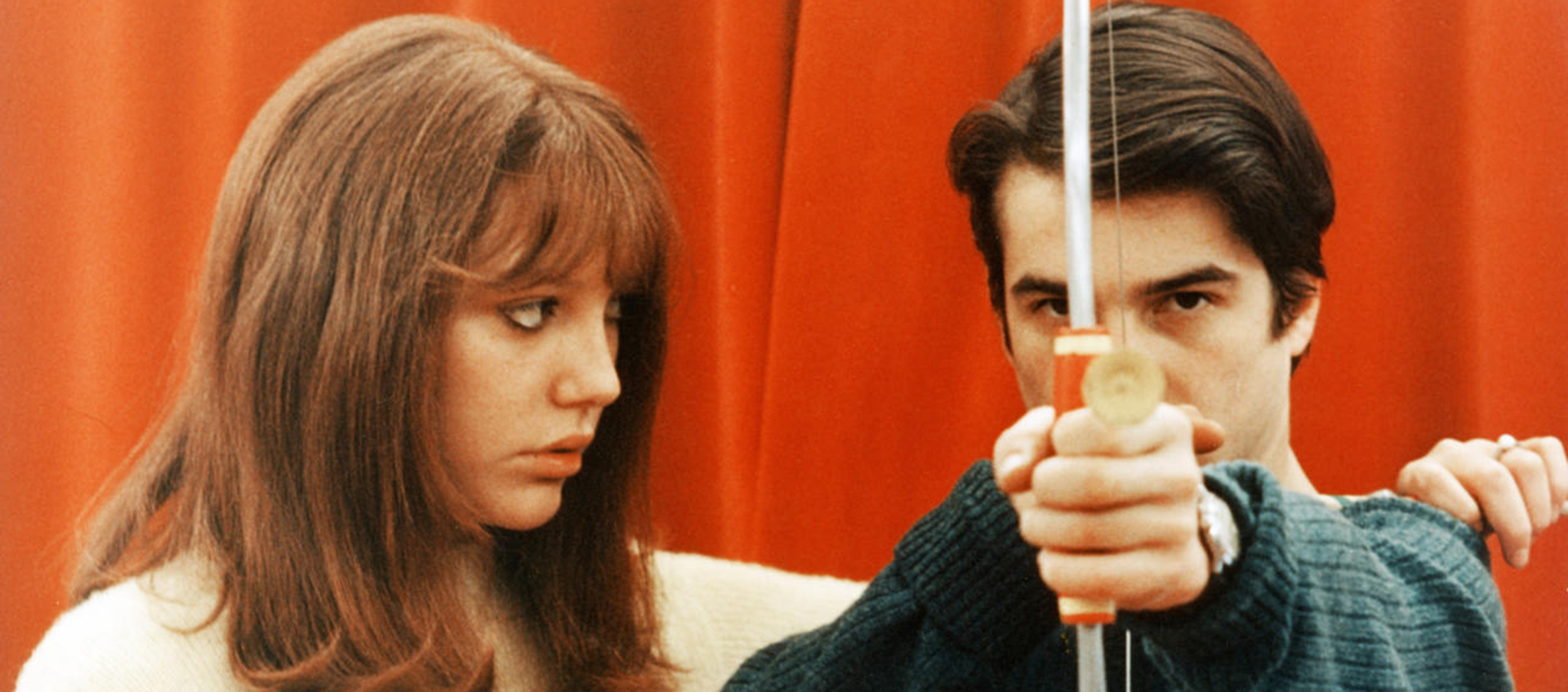 A woman is facing a man who is holding a bow and arrow outstretched in front of him.