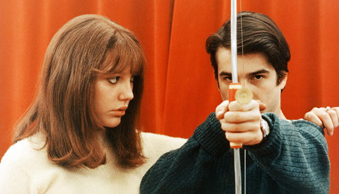 A woman is facing a man who is holding a bow and arrow outstretched in front of him.