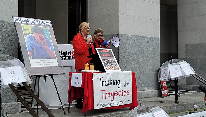 Two women stand with a megaphone behind a table with a sign stating "Tracking the tragedies."