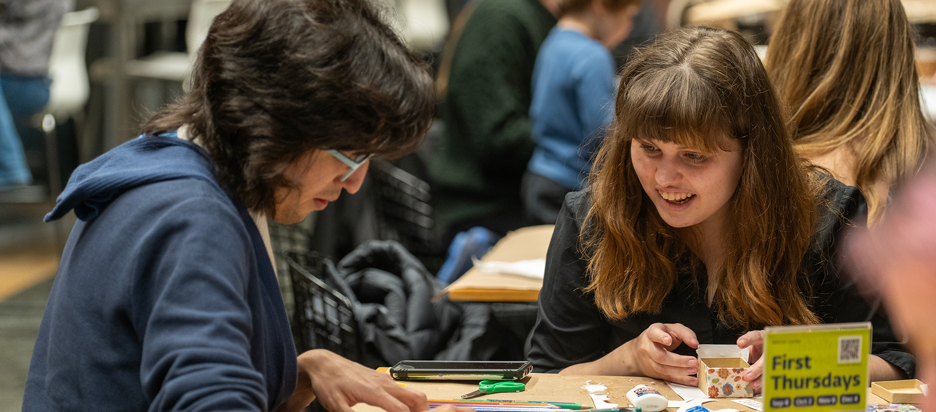 Two people sit at a table in Heirloom Café and make art. More people sit in the background.