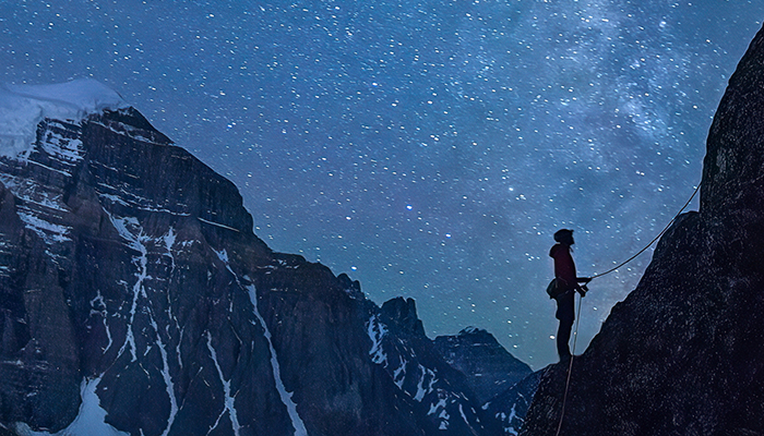The silhouette of a rock climber holding a rope in front of a mountain and starry night sky.