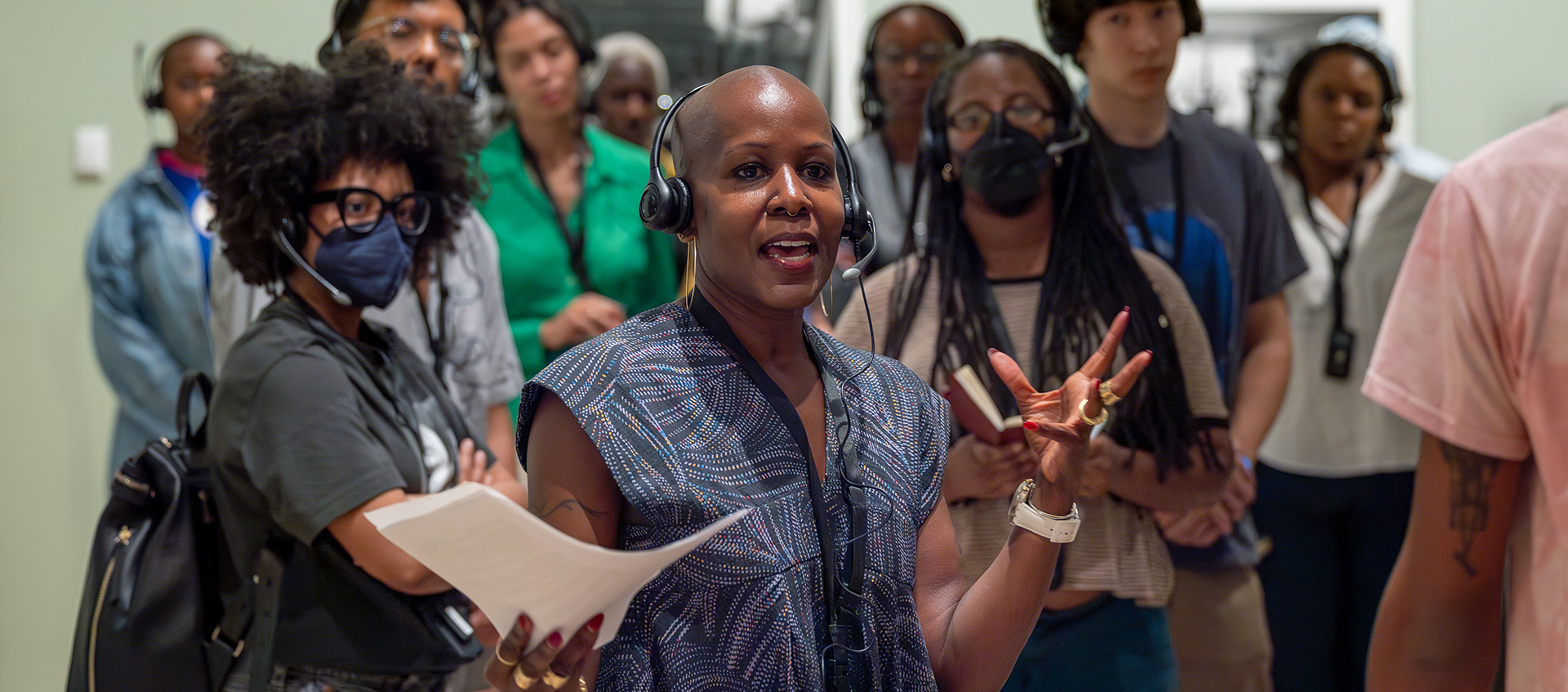 Photo of educator wearing a headset and patterned blue dress, speaking to a group of young people wearing headsets.