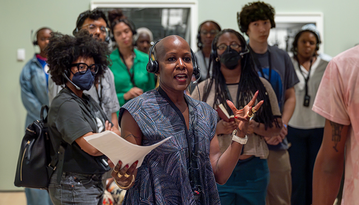 Photo of educator wearing a headset and patterned blue dress, speaking to a group of young people wearing headsets.