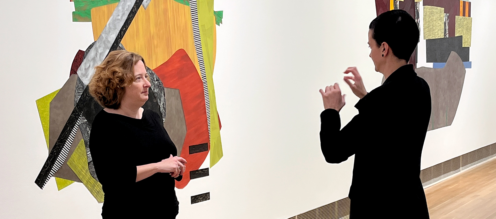 Two people stand in a gallery. The person on the right signs in ASL to the person on the left. 