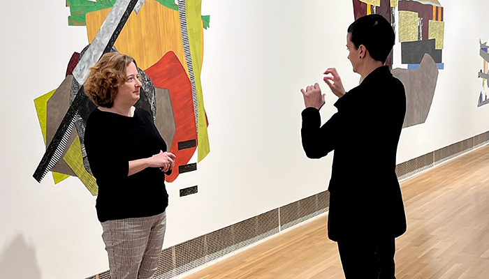 Two people stand in a gallery. The person on the right signs in ASL to the person on the left. 