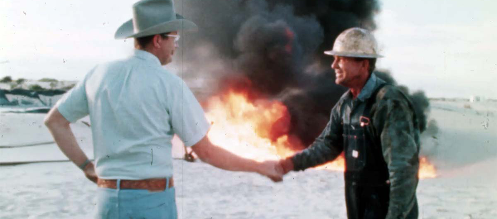A person wearing a cowboy hat shakes hands with a person in a hard hat. A blazing fire and clouds of black smoke are behind them.