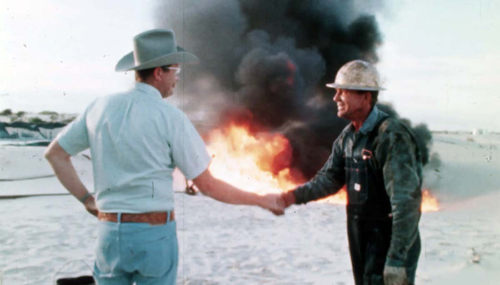 A person wearing a cowboy hat shakes hands with a person in a hard hat. A blazing fire and clouds of black smoke are behind them.
