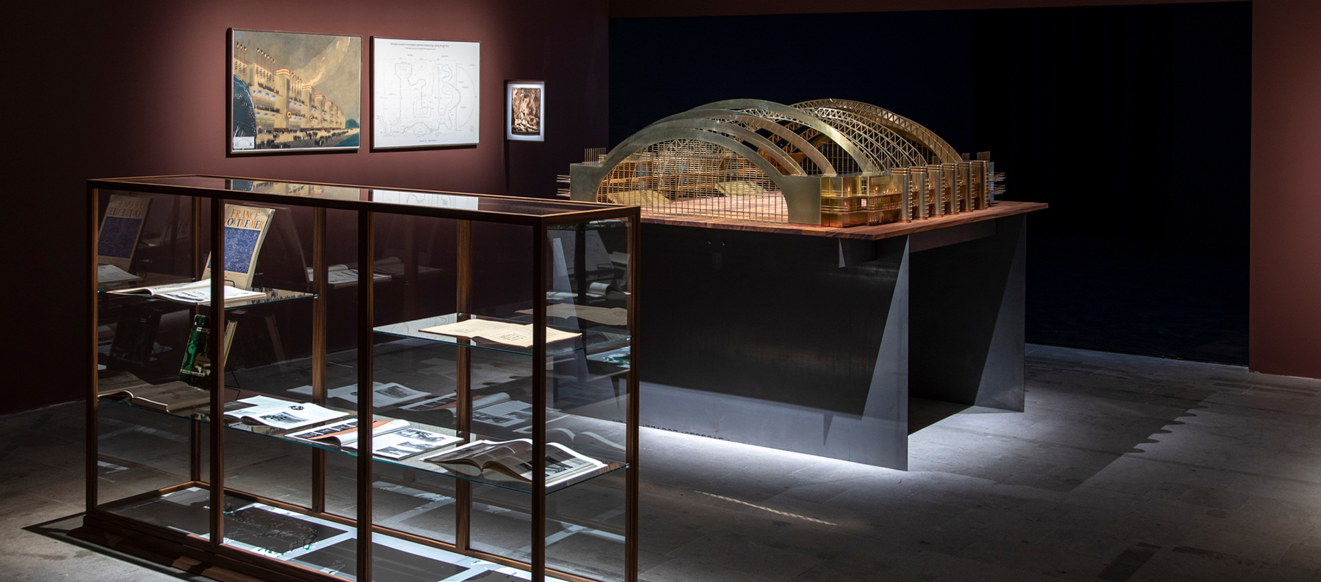 A gallery containing a glass and wood display case, an architectural model of an open structure with curved roof supports, and images of a building and building plans.  