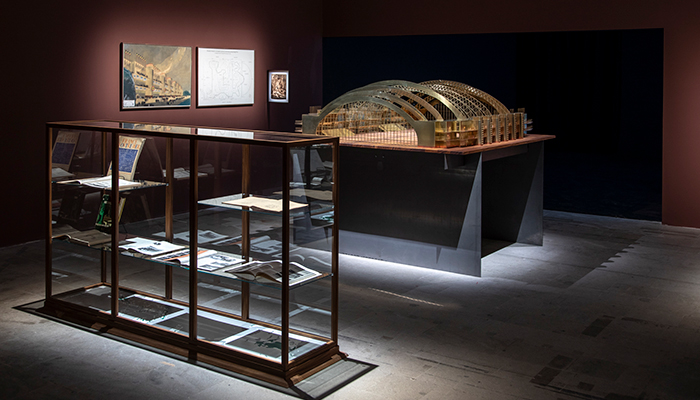 A gallery containing a glass and wood display case, an architectural model of an open structure with curved roof supports, and images of a building and building plans.  