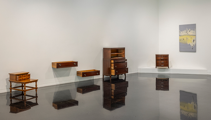 A gallery with a reflective floor displays antique furniture with partially opened doors and drawers. An abstract gray, white, and yellow painting hangs nearby. 