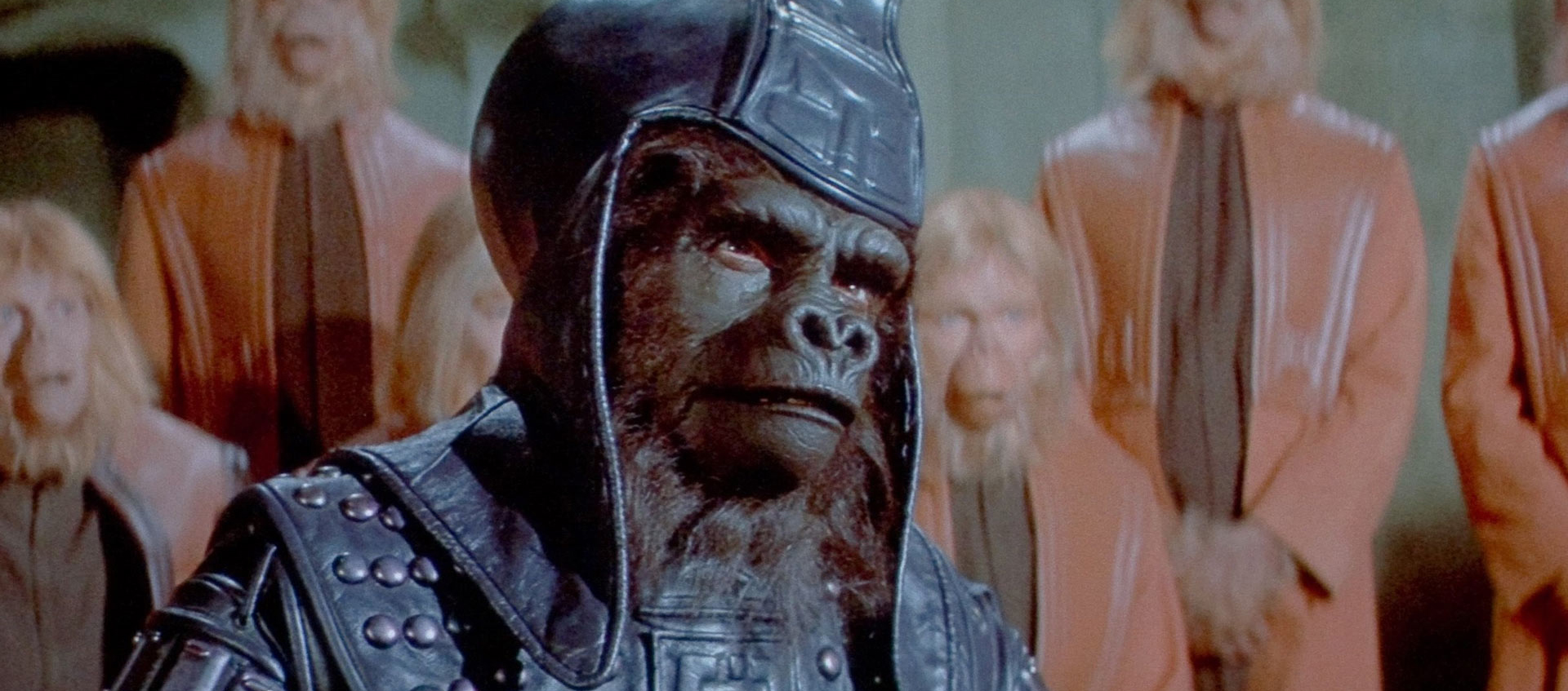 An ape wearing a leather headdress and armor stands in front of other apes wearing matching orange uniforms.