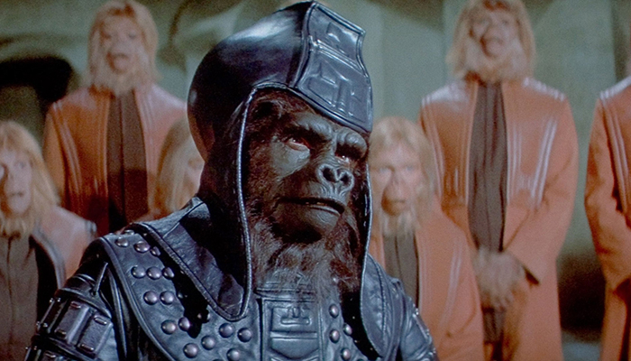 An ape wearing a leather headdress and armor stands in front of other apes wearing matching orange uniforms.