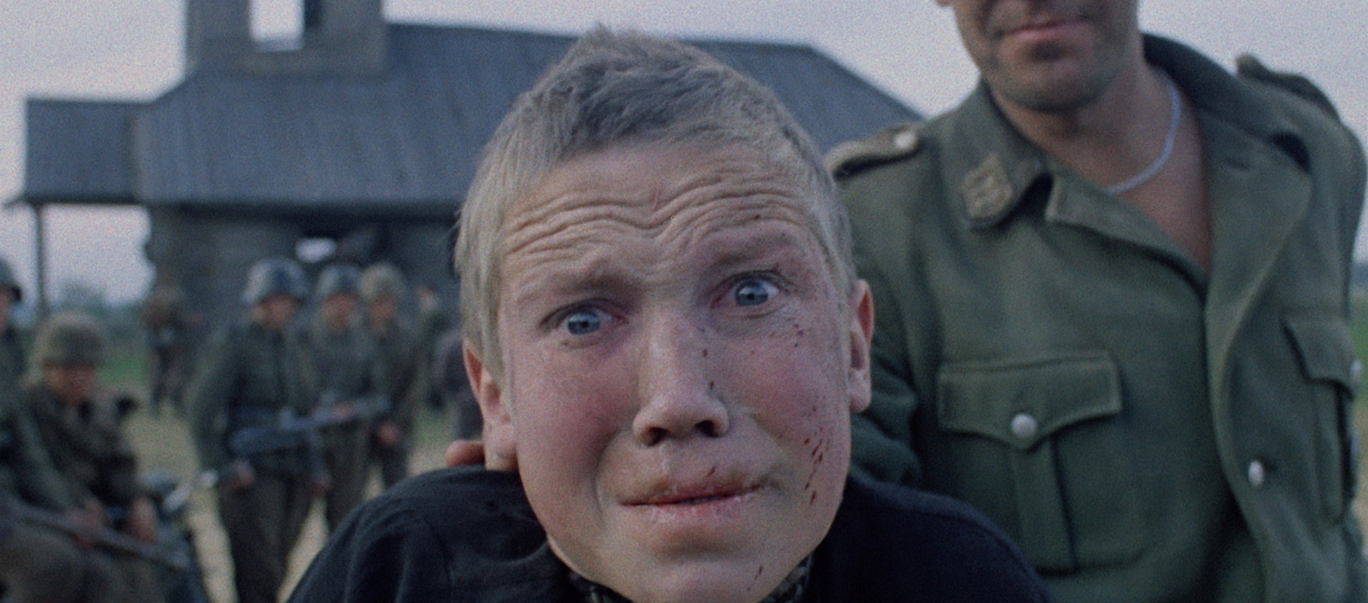 Close up on a boy's face. He is being held by a soldier with more soldiers in the background.