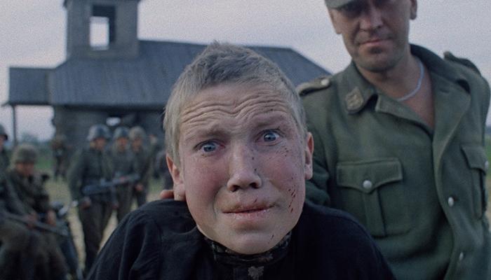 Close up on a boy's face. He is being held by a soldier with more soldiers in the background.