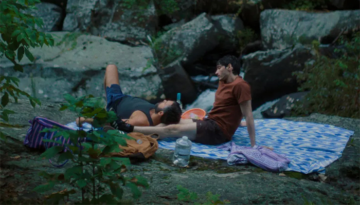 Two men sit on a blanket in the woods, one is laying with his head on the other's leg.