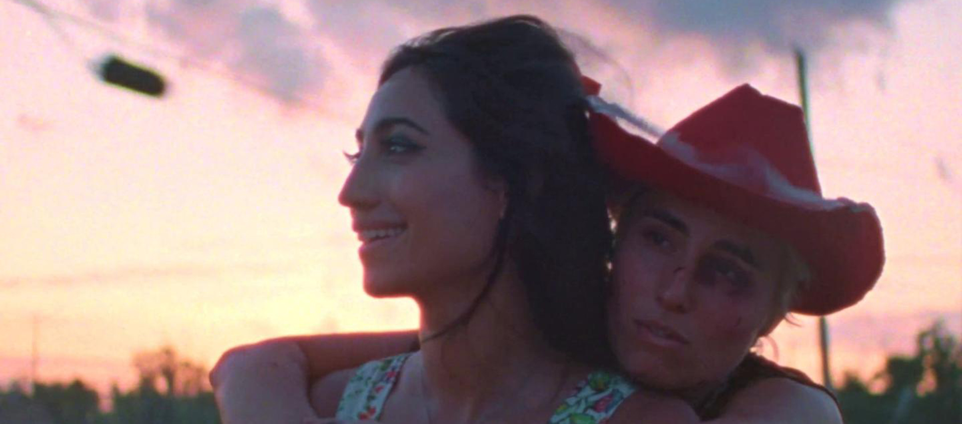 At dusk a woman wearing a red cowboy at wraps her arms around another woman's shoulders.