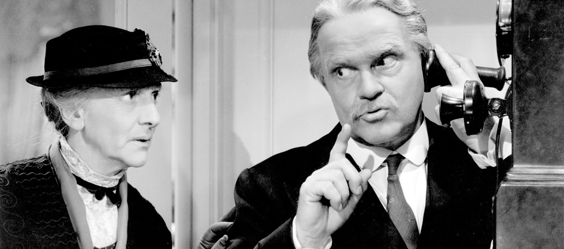 An older woman and man stand together, the man is speaking into a antique wall telephone.